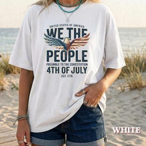 Può includere: Una t-shirt bianca a maniche corte con la grafica di un'aquila calva con le ali spiegate, colorata in rosso, bianco e blu. Il testo recita: UNITED STATES OF AMERICA WE THE PEOPLE PREAMBLE TO THE CONSTITUTION 4TH OF JULY EST. 1776.