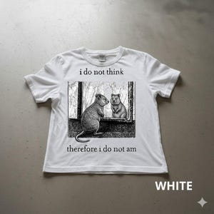 Può includere: T-shirt bianca con un'illustrazione in bianco e nero di due quokka. Uno è dentro una finestra, l'altro fuori. Il testo "i do not think, therefore i do not am" è stampato sopra e sotto l'illustrazione.