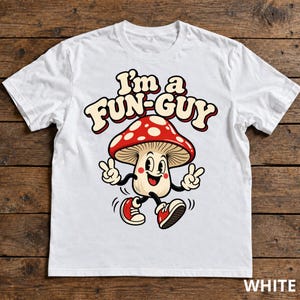 Può includere: Una t-shirt bianca a maniche corte con la grafica di un fungo dei cartoni animati con cappello rosso e pois bianchi. Il personaggio sorride, indossa scarpe da ginnastica rosse e fa il segno della pace con entrambe le mani. Il testo "I'm a FUN-GUY" è stampato sopra il fungo in un carattere retro audace.