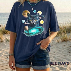Può includere: Una t-shirt blu navy con una stampa grafica di un gatto circondato da pianeti e una galassia vorticosa. Il gatto è raffigurato con occhi gialli e un anello attorno alla testa, su uno sfondo blu scuro con vari corpi celesti.