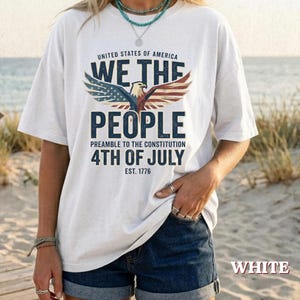 Può includere: Una t-shirt bianca a maniche corte con la grafica di un'aquila calva con le ali spiegate in rosso, bianco e blu. Il testo recita UNITED STATES OF AMERICA WE THE PEOPLE PREAMBLE TO THE CONSTITUTION 4TH OF JULY EST. 1776 in blu scuro.