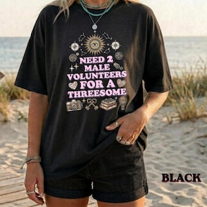 May include: A black short-sleeve t-shirt featuring a graphic print with the text NEED 2 MALE VOLUNTEERS FOR A THREESOME in pink letters. The design includes illustrations of an eye, a camera, keys, books, a hot air balloon, and stars.
