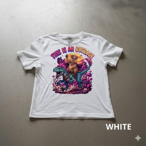 Puede incluir: Camiseta blanca con un gráfico colorido de un oso montando un dinosaurio, con el texto "THIS IS AN OUTRAGE" en rosa y morado. El diseño incluye un atardecer, setas y calaveras.