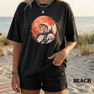 Può includere: Una t-shirt nera a maniche corte con la grafica di un leone che indossa un casco spaziale all'interno di un grande cerchio arancione. Il design include un piccolo carattere giapponese bianco sul lato sinistro del cerchio.