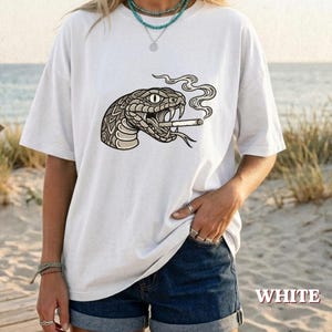 Könnte beinhalten: Ein weißes Kurzarm-T-Shirt mit einer grafischen Illustration einer Schlange mit offenem Maul, die eine Zigarette mit aufsteigendem Rauch hält. Das Schlangendesign ist in Schwarz- und Grautönen mit detaillierten Schuppen und einem Auge gehalten.