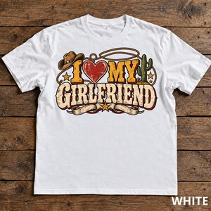 Può includere: Una t-shirt bianca con una stampa grafica che riporta il testo I LOVE MY GIRLFRIEND. Il design include un cappello da cowboy marrone, un cuore rosso, un cactus verde, un lazo e stelle gialle in un layout a tema western.