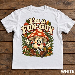 May include: A white t-shirt featuring a graphic of a cartoon mushroom character with a red cap and white spots, wearing brown boots and making peace signs. The text "I'm a FUN-GUY" is printed above the mushroom, surrounded by green ferns and autumn leaves.