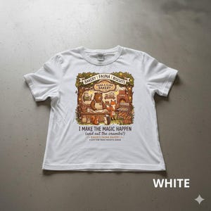 Può includere: T-shirt bianco con un'illustrazione colorata di una scena di panetteria con un orso, una volpe e altri animali. Il testo sulla maglietta recita "I MAKE THE MAGIC HAPPEN (and eat the crumbs!)" e "BAKER'S FAUNA BAKERY".