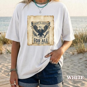 Può includere: Una t-shirt bianca a maniche corte con una grafica rettangolare raffigurante un'aquila calva con le ali spiegate, che stringe frecce e un ramoscello d'ulivo. Il testo recita Liberty & Justice For All Est. 1787 su uno sfondo color pergamena.