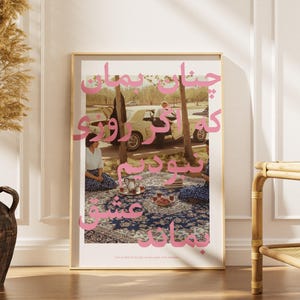 May include: A framed art print featuring a vintage car and people enjoying a picnic. The image has pink text overlayed on it. The print is displayed on a wooden floor next to a decorative vase with dried plants.
