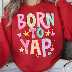 以下が含まれることがあります： 「BORN TO YAP」の文字がカラフルで遊び心のある赤いスウェットシャツ。星、蝶、足跡、花のデザインが楽しくて気まぐれな雰囲気を醸し出しています。快適でスタイリッシュなトップスです。