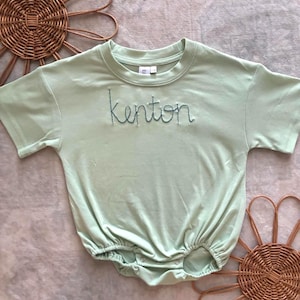 May include: A light green baby romper with short sleeves and a round neckline. The name "Kenton" is embroidered in a darker green thread across the chest. The romper has a gathered waist and snap closures at the bottom.