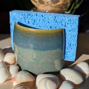 May include: A ceramic sponge holder with a blue, green, and yellow glaze, set against a blue sponge. The holder is surrounded by various seashells, creating a coastal theme. The holder is designed to hold a sponge.