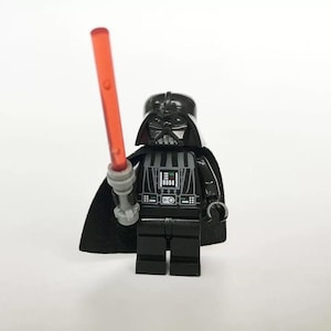 Puede incluir: Una figura de Lego negra de Darth Vader con un sable de luz rojo. La figura tiene una capa negra y una placa de pecho detallada. El sable de luz se sostiene en la mano derecha de la figura.