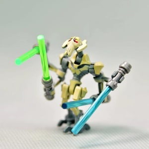 Könnte beinhalten: Eine General Grievous Actionfigur mit vier Armen, die drei Lichtschwerter hält. Die Figur ist hauptsächlich gelb und grau, mit einem weißen Kopf und grünen und blauen Lichtschwertern. Die Figur ist vor einem hellgrauen Hintergrund posiert.
