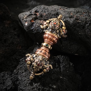 May include: Ornate, antique-style metal vajra. The vajra features a detailed design with gold and copper tones, accented by red and green gemstones. The intricate craftsmanship includes depictions of mythical creatures.