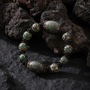 May include: A bracelet with green and cream patterned beads. The bracelet has oval and round beads, linked by black and bronze spacers. The bracelet is displayed on a dark, textured surface. The beads are patterned with concentric circles.