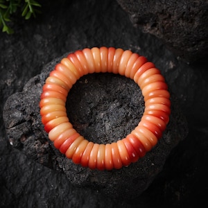 Puede incluir: Una pulsera de color coral compuesta por cuentas redondeadas y segmentadas. Las cuentas hacen una transición de un naranja pálido a un tono rojo vibrante. La pulsera se exhibe sobre una superficie oscura y texturizada, resaltando su color y forma.