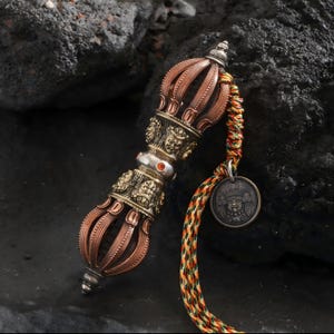 May include: Ornate bronze and copper vajra, a ritual object, with intricate carvings and a decorative handle. A colorful braided cord is attached, ending with a small, round pendant. The background is a dark, textured surface.