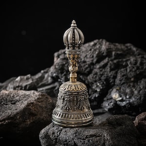 May include: An ornate silver and gold bell with intricate detailing. The bell features a crown-like top and a detailed handle with a figure. The bell is displayed on a dark, textured rock surface against a black background.