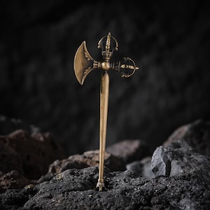 May include: A detailed, antique-style brass axe with intricate designs. The axe head features a curved blade and ornate detailing, while the handle has a slender, elongated form. The object is set against a dark, textured background, highlighting its metallic sheen.