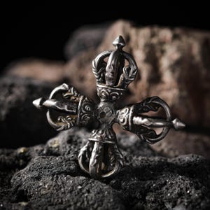 Może przedstawiać: Szczegółowy, srebrny Vajra, przedmiot rytualny, spoczywa na ciemnej, teksturowanej powierzchni. Vajra ma skomplikowane wzory i kształt krzyża z centralnym punktem i czterema promienistymi ramionami, z których każde ma element dekoracyjny.