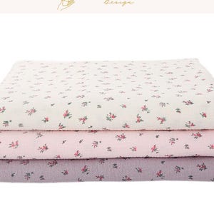 May include: Three folded fabric layers in cream, pink, and lavender, each adorned with a delicate floral pattern in red and green. The top layer is cream, the middle is pink, and the bottom is lavender. The image includes the text "EXPLOREANDSHOP Design".