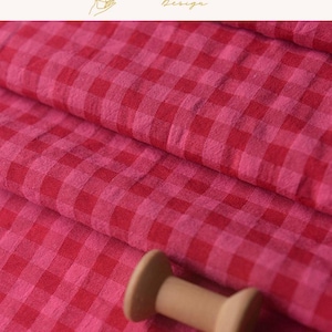 Op de afbeelding: Een gevouwen stuk stof met een levendig rood en roze vichy-ruitpatroon. Het textiel heeft een zichtbaar geweven textuur en is in zachte plooien gedrapeerd. Een kleine, lichtbruine houten klos bevindt zich op de voorgrond van de stof.