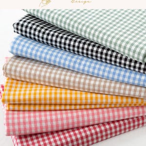 Op de afbeelding: Een stapel gevouwen stoffen met een klassiek gingham-ruitpatroon in diverse kleuren, waaronder saliegroen, zwart, lichtblauw, beige, geel, lichtroze en rood. Elk stuk stof vertoont een consistent patroon van kleine vierkante ruitjes.