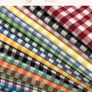 以下が含まれることがあります： 赤、黒、ピンク、黄色、青、緑、オレンジ、茶色、紫など、様々な色のクラシックなギンガムチェック柄の折り畳まれた布地が斜めに積み重なっており、上部にはEXPLOREANDSHOP Designというテキストが記載されています。