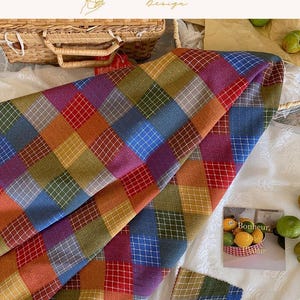 May include: A fabric featuring a multi-colored diamond pattern in shades of red, blue, yellow, green, and purple, overlaid with a white grid. The textile has a textured appearance and is displayed alongside a small card with the text Bonheur, Joie, Plaisir.