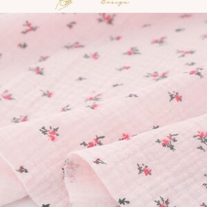May include: Close-up of a pink fabric with a delicate floral pattern. The fabric is a light pink color with small, scattered red and green floral designs. The fabric is soft and appears to be lightweight, with a slightly wrinkled texture. The text "EXPLOREANDSHOP Design" is in the top right corner.