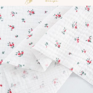 May include: White fabric with a delicate floral pattern featuring small red roses and green leaves. The fabric is folded, showcasing its texture and the scattered floral design. The top right corner has the text "EXPLOREANDSHOP Design."