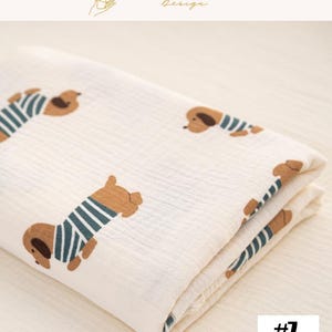 May include: Cream-colored blanket featuring a repeating pattern of brown dachshunds wearing blue and white striped shirts. The blanket is folded, showing the pattern on the top layer. The text "EXPLOREANDSHOP Design" is at the top. The number "#7" is in the bottom right corner.
