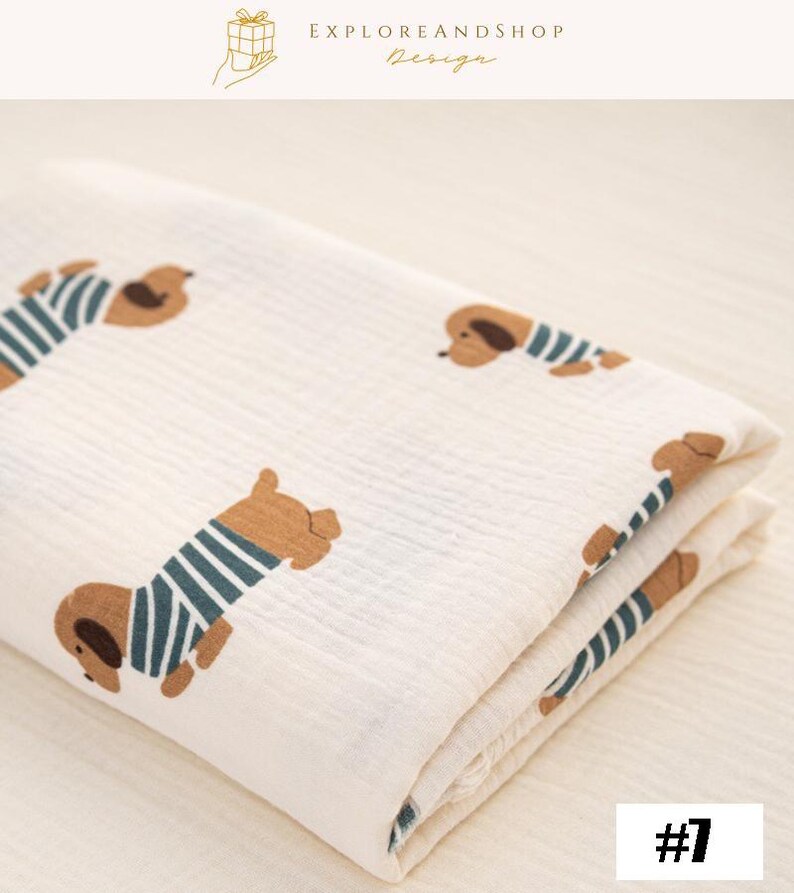 May include: Cream-colored blanket featuring a repeating pattern of brown dachshunds wearing blue and white striped shirts. The blanket is folded, showing the pattern on the top layer. The text "EXPLOREANDSHOP Design" is at the top. The number "#7" is in the bottom right corner.