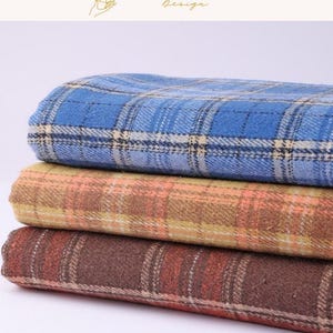 May include: A stack of three folded fabric pieces featuring a plaid pattern. The top piece is blue with white and dark blue lines. The middle piece is tan with orange and yellow lines. The bottom piece is dark brown with orange and white lines.