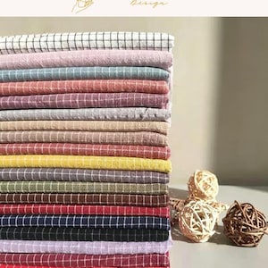May include: A vertical stack of folded fabric pieces, each featuring a grid pattern. The textiles display a variety of colors including white, pink, blue, tan, yellow, green, brown, red, black, and purple, accompanied by two woven rattan spheres.