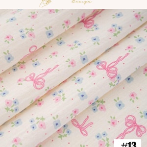 May include: Close-up of a cream-colored fabric with a delicate floral pattern. The design features small pink and blue flowers, along with pink ribbon bows. The fabric is folded, showing the texture and pattern. The number 13 is in the bottom right corner.