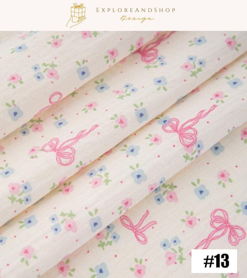 May include: Close-up of a cream-colored fabric with a delicate floral pattern. The design features small pink and blue flowers, along with pink ribbon bows. The fabric is folded, showing the texture and pattern. The number 13 is in the bottom right corner.