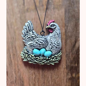 May include: A pendant necklace featuring a hen figurine with a red comb and blue eggs in a nest. The hen is white and gray with black markings. The necklace hangs on a black cord.