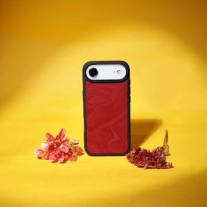 May include: A black-framed phone case with a red marble design. The phone case has a white top section with a camera lens. The case is on a yellow background with red flowers and dried plants.