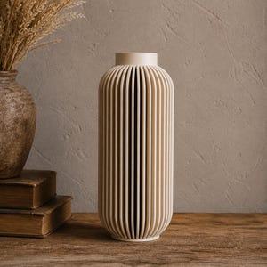 May include: A tall, cream-colored vase with vertical, ridged detailing. The vase has a rounded base and a narrow neck. The background includes a rustic wooden surface, a textured wall, and a glimpse of dried plants in a similar vase.
