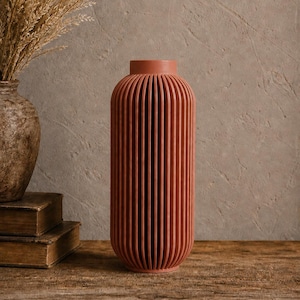 May include: A terracotta-colored vase with vertical ribbed detailing. The vase has a cylindrical body and a rounded top. The vase is displayed on a wooden surface, with a second vase and books in the background. The vase is a decorative home decor item.