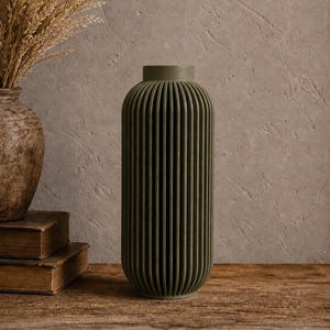May include: A tall, olive green vase with vertical ribbed detailing. The vase is cylindrical with a slightly flared top. It sits on a rustic wooden surface, with a textured wall in the background. A second, older vase and dried plants are also visible.