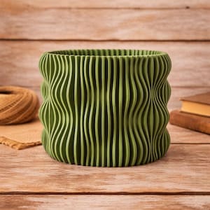 May include: A green, wavy-lined planter pot. The pot has a cylindrical shape with vertical, undulating lines that create a textured surface. The color is a muted, olive green. The pot is sitting on a wooden surface.