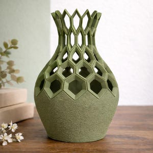 May include: A green decorative vase with a textured base and a geometric honeycomb design. The top of the vase has an openwork pattern. The vase is made of a solid material and sits on a wooden surface.
