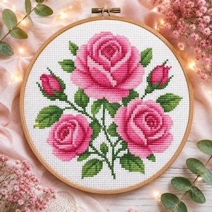 Puede incluir: Un bastidor de bordado circular de madera que contiene un patrón de punto de cruz de tres rosas rosadas y dos capullos con hojas verdes sobre un fondo de tela blanca. El diseño presenta varios tonos de rosa para los pétalos y verde para el follaje.