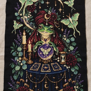 Puede incluir: Un bordado de punto de cruz sobre tela negra que representa una rana verde con turbante y túnica roja, sentada ante una mesa con una bola de cristal, cartas del tarot y un reloj de arena, rodeada de polillas verde pálido, velas y flores.