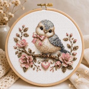 May include: A circular wooden embroidery hoop containing a cross-stitch pattern of a small blue and beige bird with a pink bow around its neck, perched on a branch surrounded by pink roses and a small pink heart, all on white fabric.