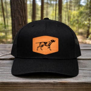 May include: Black trucker hat with a mesh back and a curved brim. The front features a rectangular patch with a brown and black dog silhouette on an orange background. The hat is sitting on a wooden surface.